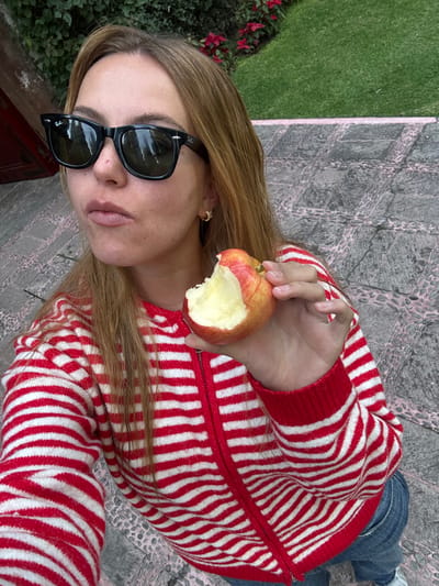 A woman with long brown hair and sunglasses holds an apple with a bite taken out. She wears a red and white striped zip-up sweater. She is standing on a concrete surface. Green grass and red flowers are visible in the background. She is wearing jeans.