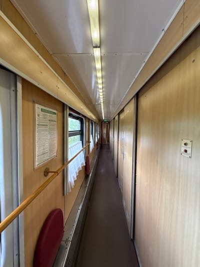 The image shows the interior of a train car, looking down the length of the aisle. On the left are white walls with linear lighting fixtures running along them. The ceiling is light wood paneled with a recessed lighting channel down the center. On the right are dark brown paneled walls. Windows with white pleated curtains are visible along the right wall, with a yellow handrail running below the windows. A sign with printed text is affixed to the wall below one of the windows. A red cushion is placed on the wall. The lighting suggests an interior setting. Based on the available information, the location within the Красненскі сельскі Савет area is a train carriage.