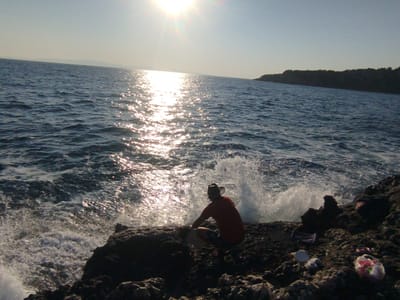A person wearing a hat and an orange shirt is sitting on rocky terrain near the sea, with waves crashing against the shore. The sun is shining brightly, reflecting off the water. In the background, a dark, tree-covered coastline is visible under a clear sky. There are also some plastic bags and other debris scattered on the rocks. The scene is set in Karaburun, Turkey.