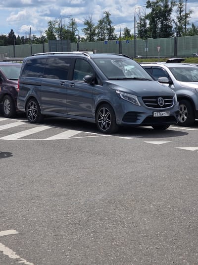 A grey Mercedes-Benz V-Class van, license plate T776KC 799, is parked in a parking lot in Sholokhovo, Russia, under a cloudy sky. The van is positioned behind a maroon vehicle and next to a silver SUV. The parking lot is paved with asphalt and marked with white lines. A sound barrier covered in green panels is visible in the background, along with trees and a lamppost.