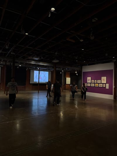 People walk through a large, dimly lit warehouse-style space with exposed ceiling structures. On one side, a purple wall displays framed artwork. A projection screen shows a cartoonish diagram, attracting a small group. The polished concrete floor reflects the sparse lighting. One individual stands apart, closer to the viewer, facing away.