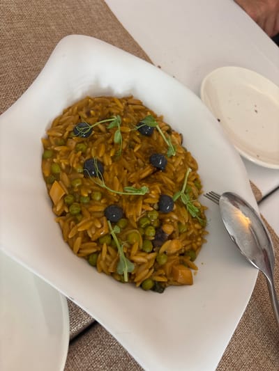 A close-up, overhead shot captures a white, uniquely shaped plate filled with a portion of orzo pasta dish. The pasta is coated in a rich, golden-brown sauce and mixed with vibrant green peas and what appear to be small pieces of mushroom or other protein. Three dark olives are strategically placed on top of the pasta, and delicate green microgreens garnish the dish. The plate rests on a textured, brown placemat. To the right of the plate, a silver spoon and fork are visible, suggesting a meal is being served or consumed. The background is blurred, but hints of white tableware and a person's hand indicate a dining setting. The lighting is natural and soft, highlighting the textures and colors of the food. This scene is likely from a restaurant in Carboneras, Spain, given the provided context.