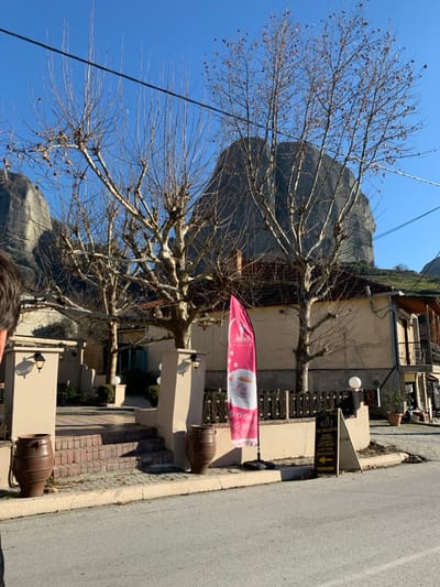 The image, taken in Kastraki, Greece, presents a sunlit street scene with prominent rock formations in the background. On the right, an asphalt road runs parallel to a brick-patterned sidewalk. To the left, buildings with light-colored facades line the street, featuring outdoor seating areas with wooden tables and chairs.
Several mature, leafless deciduous trees, likely pollarded, stand along the sidewalk, their intricate branch structures reaching towards a clear blue sky. Overhead, thin power lines traverse the frame. Behind the buildings and trees, immense grey rock pillars, characteristic of the Meteora region, dominate the landscape under the bright, clear sky.
Decorative large, antique-style clay pots (amphorae) are placed on elevated stone bases in front of the buildings. A bright pink flag banner, featuring a coffee cup logo and the word "COFFEE" (possibly "JIMBO'S" at the top), is visible. Nearby, a black and white A-frame sign advertises "G.B.P." and services including "BREAKFAST, LUNCH, COFFEE, COCKTAIL, INTERNET," with an arrow indicating "PARKING." The top of a person's head is visible at the very bottom of the frame. The scene suggests a pleasant day in late winter or early spring in a scenic village setting.