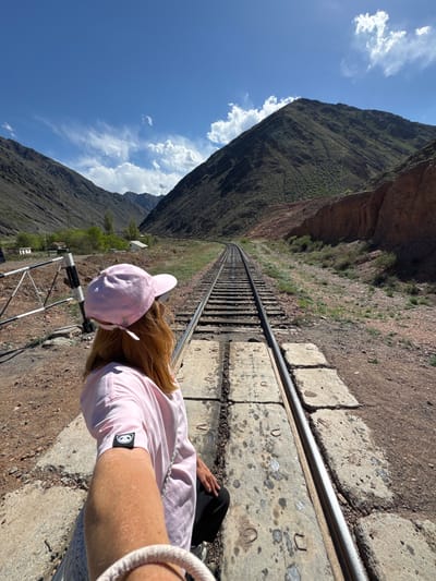 A selfie, shot from the side, depicts a person with long blonde hair wearing a pink cap and a pink shirt, standing on train tracks. The scene is an outdoor environment, likely a mountain pass, with a railway track running between the base of two mountains. The mountains rise dramatically on either side, meeting a blue sky with scattered clouds. The train tracks are set on concrete blocks, and the rails converge in the distance, indicating the curvature of the track. Some buildings are visible at the bottom of the mountains, and a black and white gate stands beside the person. The location is identified as Krasnyy Most, Kyrgyzstan.