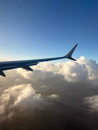 The image shows an airplane wing in flight. The wing is gray and has several flaps extended. The background is a clear blue sky on the left and white clouds on the right. The location is Unknown, United States of America.