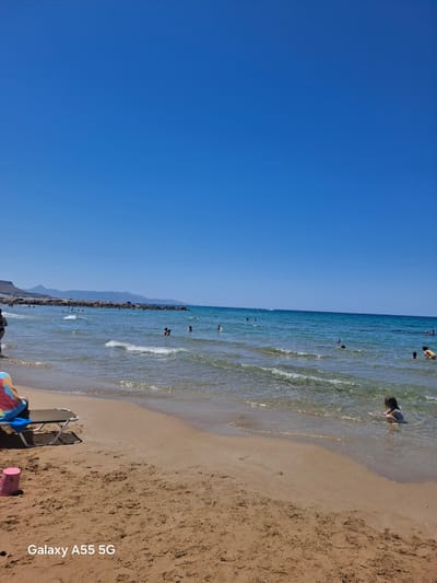 This image captures a bright, sunny beach scene in Gouves Municipal Unit, Greece, under a clear, cloudless azure sky. The foreground shows light brown sand with visible textures and footprints. On the left, a folding beach chair with blue and multicolored fabric rests near the water's edge, adjacent to a pink plastic bucket or cup. The bottom left corner of the image is watermarked with "Galaxy A55 5G".

The midground features clear, turquoise-blue water with small, gentle waves breaking on the shore. Multiple individuals, including both adults and children, are distributed throughout the shallow and mid-depth water. On the far right, a small child with dark hair is seen wading, submerged to the waist. Other figures are swimming or wading further out, with one person in dark swimwear partially visible on the far left.

In the background, a rocky formation or breakwater extends into the sea from the left, behind which a range of distant mountains with a faint blue tint rises against the horizon. The deeper sea maintains a vibrant blue color, meeting the clear sky at a defined horizon line.