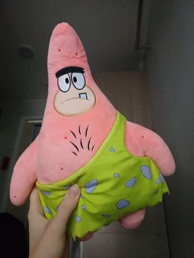 A human hand, exhibiting light skin and visible fingernails, grasps a pink plush toy of Patrick Star, depicted in a primitive style. The toy features an elongated, conical head, an embroidered face with wide eyes, a thick black unibrow, an open mouth revealing a single square upper tooth, and several short, black, stitched lines on its chest resembling hair. It is dressed in a light green fabric garment patterned with irregular light blue spots, resembling a loincloth. The hand holds the lower-right side of the plush, engaging with the green garment. The background is an out-of-focus indoor environment located in 서대문구, South Korea. The background consists of a dark gray wall, with a circular recessed light fixture visible in the upper-left ceiling and a vertical, lighter-toned architectural element on the right side.