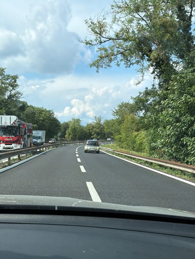 The image shows a road in Basaldella / Basandiele, Italy. The road is paved and has white lane markings. There are several vehicles on the road, including cars and trucks. Some of the trucks appear to be emergency vehicles, with one red and white truck that looks like a fire engine. The scene is bordered by trees and foliage on the left and a guardrail on the right. The sky is partly cloudy. The overall impression is of a traffic jam or a stopped procession of vehicles, possibly due to an emergency. The time of day appears to be daytime, and the weather is fair. No people are clearly visible, but the presence of emergency vehicles suggests a possible event requiring their attendance. Some text is visible on the side of the fire engine, including "MAGIRUS" and "IVECO". There also appears to be some text on the side of another truck that is difficult to read. The image is taken from a vehicle, looking forward along the road.