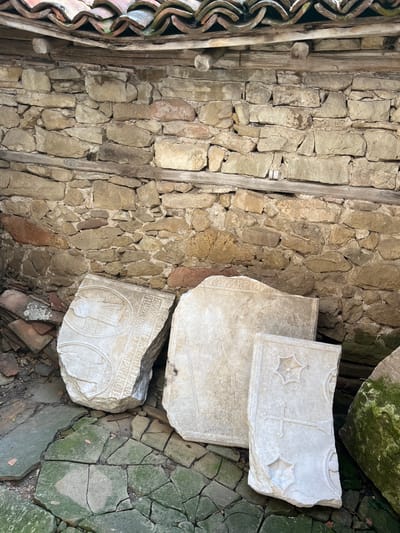 In Zheravna, Bulgaria, an outdoor scene depicts fragments of carved stone slabs leaning against a weathered, rough-hewn stone wall. The wall is constructed from irregular, tan and reddish-brown stones, with horizontal wooden beams reinforcing the structure. Above the wall, a section of a curved, tiled roof, possibly from an old building, is visible, with some tiles showing signs of age and moss growth.
In the foreground, several large, flat pieces of stone are arranged on a cracked, green-tiled surface that appears to be a floor or pavement. The stone fragments are intricately carved. One large slab, on the left, features circular engravings with figures and text in an ancient script. Another slab, to its right, is largely plain but has some faint text at the top. A smaller, upright slab on the right displays a cross and decorative star-like motifs. In the lower right corner, a large, moss-covered stone, possibly another fragment, is partially visible. The ground around the slabs is littered with smaller stone pieces and debris. The overall impression is of an archaeological site or a collection of historical artifacts stored outdoors.