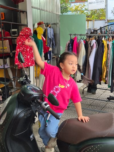 The image depicts an outdoor commercial scene in Hội An Đông Ward, Vietnam. A young Asian female child, estimated 3-5 years old, is positioned between the handlebars and seat of a dark-colored Honda scooter parked on the left. She wears a pink short-sleeved t-shirt with a light-colored graphic, light blue pants, and light-colored slip-on clogs, with her dark hair styled in two pigtails and a silver-toned necklace with a pendant. Her left hand rests on the textured brown scooter seat, while her right arm is raised and her hand open; she looks towards the right with a slightly open mouth and an expression of curiosity. The scooter features a dashboard and rearview mirrors.

Behind the child and scooter, two metal clothing racks are heavily laden with various garments, including shirts, jackets, and other apparel in multiple colors. Partially visible headless mannequins display clothing items behind the leftmost rack. The immediate ground is a light-colored, patterned paved surface. The background consists of corrugated metal siding on the left, a light green panel, and trees. A white sign reading "PAPAYA GARDEN HOMESTAY" is visible in the upper right. The lower legs of an adult are partially discernible behind the rightmost clothing rack.
