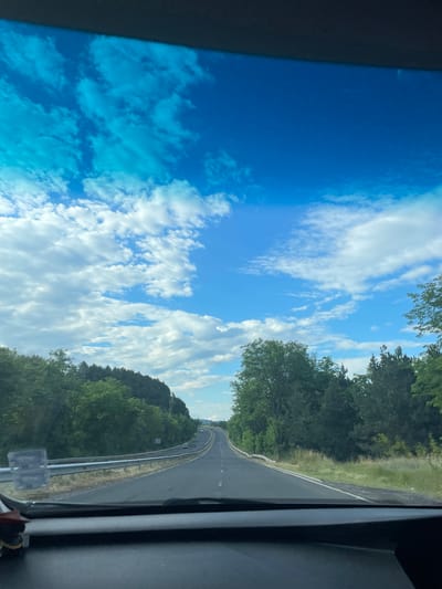 The image shows a view from inside a vehicle, looking out through the windshield. The road ahead is asphalt, with white lines marking the lanes. The road curves to the right and disappears into the distance. On either side of the road, there are trees and vegetation. In the background, there are hills covered in trees. The sky is blue with scattered white clouds. The scene appears to be a rural or suburban area, possibly outside of Balabanovo, Bulgaria, given the context. There are no people or specific objects that would strongly indicate a precise location within the city. The image captures a typical road view during daytime.