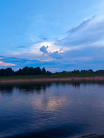 The image depicts a serene natural landscape at dusk, likely in the vicinity of Vorogovo, Russia, given the presence of a wide river and a densely forested bank. The sky is a vibrant gradient of blues and purples, with scattered clouds catching the last rays of sunlight. A prominent cloud formation in the upper right quadrant resembles an animal, adding a touch of whimsy to the scene.
The river occupies the foreground, its surface reflecting the colors of the sky and the distant trees. Gentle ripples suggest a calm atmosphere. The middle ground features a sandy riverbank, transitioning into a thick band of dark green trees that form a natural barrier along the water's edge. Beyond the trees, on a slightly elevated plain, are scattered buildings indicative of a rural settlement, with some power lines visible. The overall impression is one of peaceful tranquility, characteristic of a rural evening by the river. There are no people or animals visible in the frame.