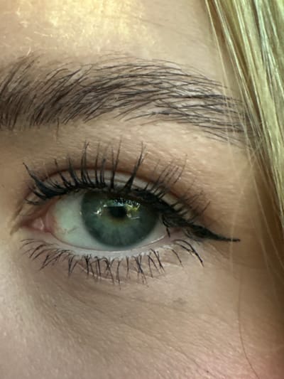 The image depicts a close-up of a human eye, positioned on the right side of the frame. The eye has long, dark eyelashes, and a thin line of black eyeliner is visible along the upper eyelid. The eyebrow, located above the eye, is thick and light brown in color. The iris appears to be a mix of green and blue, with some lighter patches. The surrounding skin is light-toned with visible pores and fine lines. Blond hair is visible at the top left. There is no information in the image to suggest a location in the city of Casetta, Italy.