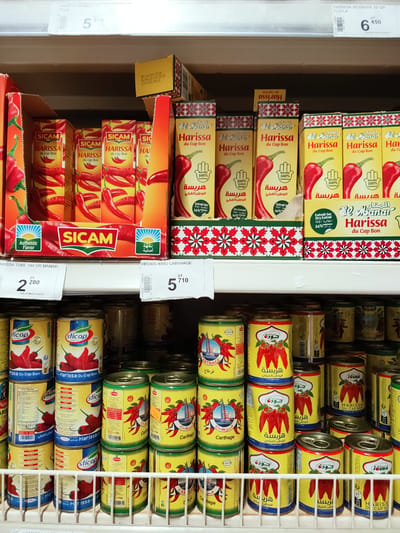 This media file shows a close-up, eye-level view of a retail shelf stocked with various brands and sizes of harissa, a spicy paste common in North African cuisine. The products are primarily in cans and boxes, displaying vibrant imagery of red chili peppers. The dominant colors are red, yellow, and green, reflecting the ingredients and branding.
The scene is set in a supermarket or grocery store, evidenced by the shelving and product arrangement. The lighting is bright and artificial, typical of an indoor retail environment. The focus is on the products themselves, with no people visible.
Several brands are present, including "SICAM," "El Manar," "sticap," and "JOUDA." Text on the packaging is predominantly in Arabic and French, with some English as well. Visible text includes product names like "HARISSA," "du Cap Bon," and brand names. Nutritional information and product details, such as weight and origin, are also visible on some packages. Prices are displayed on some items, for example, "2 280 DT" and "5 710 DT". The presence of expiration dates, such as "DF124/03/2026" and "DL00123/03/2029," indicates that the products are for sale. The overall impression is one of abundance and variety within a specific product category.