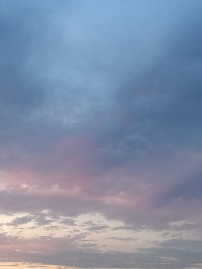 A vertical, high-angle photograph captures a sky filled with clouds during either sunrise or sunset. The upper portion of the frame is dominated by a pale blue expanse, transitioning into darker blues and grays as it descends. A band of soft pink and purple hues appears in the lower-middle section, suggesting the influence of the sun's light on the clouds. The bottom of the image shows lighter, thinner clouds tinged with a yellowish-white color, indicating the horizon. The image does not contain any discernible urban features or any individuals, suggesting it was taken in an open area. The location is Kestřany, Czechia.