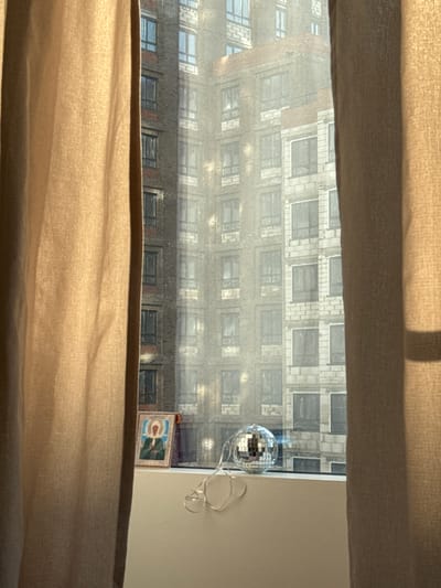 The image shows a partial view from a window covered by a light-colored curtain. Through the opening in the curtains, a building across the street is visible, featuring a brick facade and a grid of windows. Inside the room, adjacent to the window, there's a disco ball hanging with a wire. A small framed picture is situated next to the window as well. There are no visible people, actions, or interactions. The location, Люк, Russia, is not ascertainable from the image.