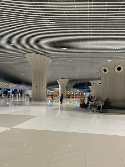 The image depicts the interior of a modern airport terminal. The architecture features a high, curved ceiling with a repetitive slatted design and large, conical columns that support the structure. Several people are visible in the scene, some walking, others sitting on benches, and some appearing to be waiting in line at check-in counters in the background. Luggage carts and benches are also present. The flooring is highly polished and reflective, creating mirrored images of the ceiling and columns. The overall impression is one of a spacious, contemporary public space. This airport is located in Baji Mangngai, Indonesia.