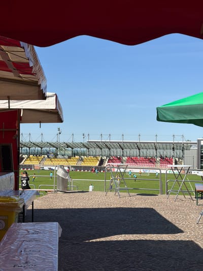 The image shows a view of a soccer field with red and yellow seating visible in the stadium. The soccer field appears to be located in Fentange, Luxembourg. The perspective is from behind a vendor's area, with a red awning on the left, and a green awning visible above the field. A cobblestone walkway is on the right, in shadow. There are people visible on the field, and it appears to be a game or practice in progress. There is a garbage can and what looks like packaged items in the foreground. The sky is blue and clear.