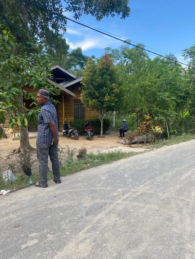 A man stands in Gampongaceh, Indonesia, next to a rural road. He wears a patterned shirt, dark pants, and a cap. Behind him is a small wooden house and two parked motorbikes. Trees and vegetation surround the buildings and the road. Another individual stands further down the road, near a small structure. The sky above is blue with some clouds.