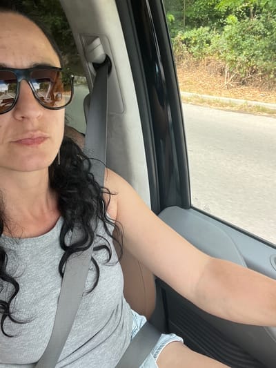 In an indoor, likely vehicle, setting, a woman with dark curly hair and sunglasses is captured in a selfie. She is wearing a grey ribbed tank top and light-wash denim shorts. A grey seatbelt is fastened across her chest. The background, visible through a window, suggests an outdoor environment with greenery and a road. The image does not provide specific clues to confirm the location within Khust, Ukraine.