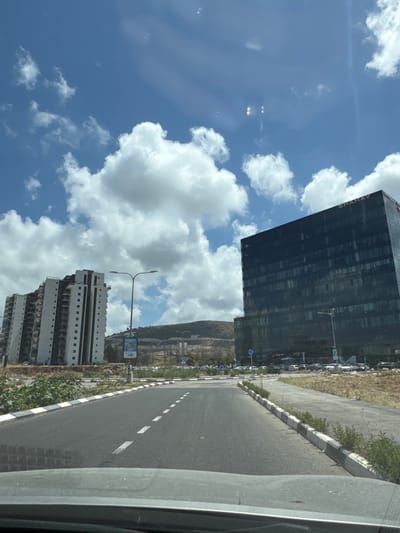 The image shows a street scene in Tirat HaCarmel, Israel, on a sunny day with scattered clouds. In the foreground, a dark-colored car's dashboard is visible, indicating the photo was taken from inside a vehicle. The road ahead is asphalt with white lane markings.
On the left side of the road, a tall, modern residential building with a white and gray facade is seen. It has multiple floors with balconies and dark-colored window sections. Adjacent to it, a lamppost stands tall. Further down the road, there's a billboard.
On the right side of the road, a sleek, dark glass-fronted office building dominates the scene. The word "SHAHAV" is visible in white lettering on its upper facade. This building appears to be taller and more contemporary than the residential building. In front of the office building, a parking lot with several cars is visible.
The background consists of a hillside with sparse vegetation and some construction or infrastructure work. The sky is a bright blue with fluffy white cumulus clouds. The overall impression is of a developing urban or suburban area in Israel.