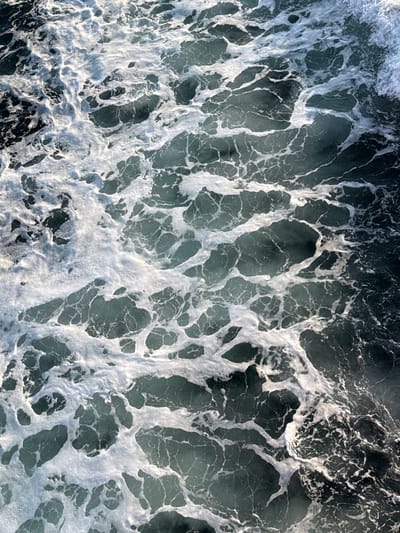 The image displays an overhead perspective of a turbulent body of water, consistent with the coastal environment of Palaiopoli, Greece. The scene is dominated by intricate patterns of white sea foam coexisting with dark teal-blue water. The foam, varying in density from thick, frothy aggregations to delicate, interconnected threads, covers much of the surface. Strong, direct illumination creates distinct highlights on the foam's surface, while shadows reveal its three-dimensional texture. Patches of darker, relatively clearer water are visible between the foamy structures, suggesting underlying currents or wave action. No human figures, objects, or structures are present.