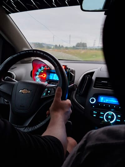 A first-person perspective shot from inside a car driving on a rural road towards Simakovo, Russia. The driver's hands are on the steering wheel and their right thumb is visible. The car's dashboard displays the speedometer, fuel gauge, and infotainment system. The windshield has cracks. The sky is overcast. The surrounding landscape consists of trees and power lines.