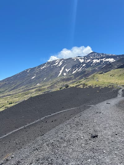 The image shows a view of Mount Etna, a stratovolcano located near Linguaglossa, Italy. In the foreground, a steep slope of dark volcanic ash and gravel dominates the scene, with a walking path winding through it. In the middle ground, the terrain transitions to green grassy slopes dotted with patches of vegetation. Further up the mountain, the dark volcanic rock is interspersed with streaks of white snow, even on this sunny day. The summit of Mount Etna is visible in the background, with a plume of white smoke or steam rising from it, indicating volcanic activity. The sky is a clear, vibrant blue with a few wispy clouds. A small group of people can be seen walking on the grassy slope in the middle ground, providing a sense of scale to the vast landscape.
