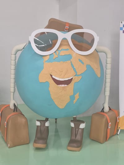 The image depicts a whimsical, anthropomorphic globe figure dressed for travel. The globe, representing Earth, has a smiling face and is wearing large, white-framed sunglasses and a brown hat. It has segmented, tube-like arms and legs. Attached to each hand is a brown suitcase with orange straps. The figure stands on two brown boots with white tops. The scene appears to be indoors, with a light-colored wall in the background and a green floor. There are no people visible in the image. The globe figure is positioned centrally in the frame, facing forward. The overall impression is of a playful and engaging display, possibly part of a larger exhibit or public art installation. There is no visual information to suggest a location in Proshyan, Armenia.