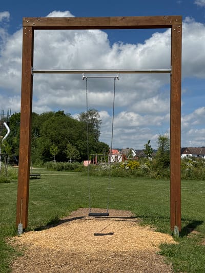 An outdoor swing set made of dark brown wooden beams and a silver metal bar stands on a grassy area with a wood chip base. A single black swing seat hangs from metal chains. In the background, there is a line of trees, houses, and a cloudy blue sky.