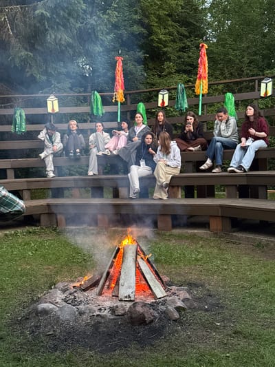 In Ratnieki, Latvia, a group of young people are gathered on a wooden tiered seating area, facing a campfire in the foreground. The campfire is actively burning, with visible flames and smoke rising. The seating area is constructed from dark wood and is surrounded by green grass and trees, indicating an outdoor setting, likely a park or camping ground. Several people, appearing to be teenagers or young adults, are seated on the tiers, dressed in casual clothing. Some are looking towards the campfire, while others are engaged in conversation or looking at their phones. The atmosphere appears relaxed and social. Decorative elements, such as colorful fabric streamers and lanterns, are visible along the top of the seating structure, adding to the ambiance. The image captures a moment of communal gathering around a fire, likely during an evening event.
