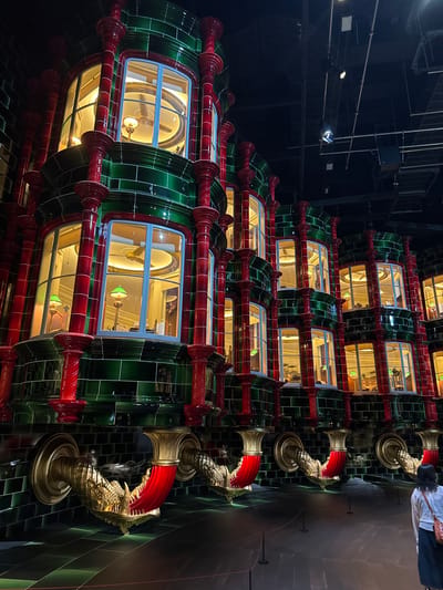 The image shows a multi-story building facade with a distinctive architectural style, featuring several stacked modules. Each module is adorned with green and red tiles, and has large windows revealing interiors lit with warm light. Within the windows, objects resembling lamps and other decorations can be discerned. To the right of the building facade are ornate, golden dragon-shaped structures, possibly decorative elements or drain spouts. The environment suggests an indoor setting, possibly a theme park or exhibit. The presence of an individual partially visible in the upper right corner indicates the scale of the structure. While the location in Nerima, Japan, is mentioned, the image does not explicitly confirm that information.
