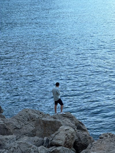 A lone man stands on a rocky outcrop overlooking a vast body of blue water. The man is viewed from behind, wearing a light-colored shirt and dark shorts. The rocks are rough and textured, characteristic of a coastal or seaside environment, likely on the coast of Baunei, Italy. The water is rippled, suggesting a gentle breeze. The overall scene evokes a sense of contemplation or solitude in a natural setting.