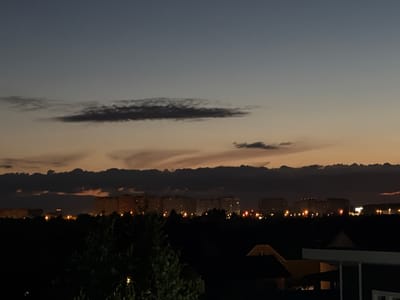 The image captures a twilight scene over the city of Martemyanovo, Russia. The sky transitions from a deep blue at the top to a soft orange and pale yellow near the horizon, with layered clouds present in the upper and middle sections of the sky. In the distance, a cluster of multi-story residential buildings is visible, illuminated by interior lights and streetlights. These buildings form a horizontal line across the frame, punctuated by small, bright lights. In the foreground, a dark, silhouetted landscape of trees and rooftops dominates the lower portion of the image. The overall impression is of a quiet evening cityscape.