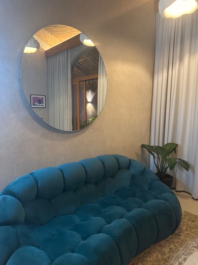 The scene depicts an interior space, consistent with a modern lounge or guest area in El Nido, Philippines. In the foreground and midground, a large, curving sofa upholstered in teal velvet features a distinctive bubble or modular tufted design. The walls are finished with a textured, light gray, concrete-like material. A large, circular, frameless mirror is mounted on the left wall above the sofa.
The mirror's reflection reveals a high ceiling section with woven natural material and a prominent wooden beam, along with two modern, white, cloud-shaped pendant light fixtures. Also visible in the reflection is a dark wood-framed doorway leading to an outdoor or semi-outdoor area featuring a thatched roof detail, a wall-mounted up-and-down light, and green foliage. A framed picture, depicting a person's lower legs in dark boots standing in a purple puddle, is reflected on an opposing wall.
To the right of the sofa, floor-to-ceiling light-colored curtains obscure a window or doorway. A potted plant with broad, dark green leaves sits on the floor between the sofa and the curtains. A patterned rug in muted tones is partially visible under the right side of the sofa. No individuals are present, and no actions or interactions are depicted.