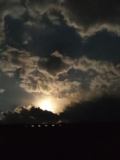 The image depicts a night sky with a full moon partially obscured by dramatic, textured clouds. The moon is the primary light source, casting a bright, golden glow through the gaps in the clouds, illuminating them from behind with a luminous halo. The clouds themselves are a mix of dark, heavy masses and lighter, more wispy formations, creating a sense of depth and movement. The overall effect is one of a powerful and somewhat brooding celestial display.
Below the sky, the silhouette of a dark landscape is visible. It appears to be a rooftop or a similar elevated structure, with a series of evenly spaced, illuminated shapes along its edge. These shapes are oval and appear to be reflecting or emitting a soft light, possibly from lights on the structure itself or from something behind it. The darkness of the foreground contrasts sharply with the brightness of the moon and illuminated clouds.
The time of day is clearly night. The weather appears to be cloudy, with the clouds playing a significant role in the composition of the sky. The location context of Qarghaly, Kazakhstan, suggests a potentially rural or suburban setting, though no specific identifiable landmarks are present in the image to confirm this. The image evokes a sense of awe and perhaps a touch of mystery due to the dramatic lighting and the obscured moon. No visible text is present in the image.