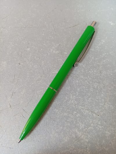 A bright green ballpoint pen is positioned diagonally on a textured gray surface. The pen has a silver clip and a silver tip. The surface appears to be made of a rough material, possibly concrete or a similar textured flooring, with visible scratches and speckles. The lighting suggests it's an indoor environment, but the specific location in Rezina, Moldova, is not discernible from the visual information provided. There are no people or actions depicted in the image.