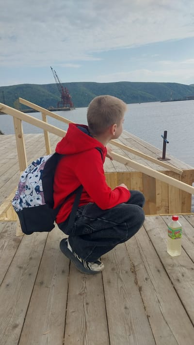 A young boy, appearing to be pre-teen, is depicted in a deep squat on a wooden plank pier in Nikolayevsk-on-Amur, Russia. He wears a red hooded sweatshirt, dark jeans, and dark athletic shoes with white markings. A backpack with a white background and numerous cartoon cat faces in various colors (black, grey, purple, pink) is slung over his left shoulder, with a solid black base. His head is turned to the right, and his gaze is directed towards the expansive body of water (likely the Amur River) and the distant landscape.

To the right of the boy, a set of light-colored wooden railings from a descending structure (steps or ramp) is visible. A plastic bottle with a red cap and a partially visible green label (displaying "Hoops Hoops") containing a clear, light-colored liquid rests on the wooden planks near his right foot.

In the middle ground, the broad river extends towards the horizon. The distant shoreline is characterized by rolling hills covered with dense green forestry. Prominently positioned on the distant left bank are two large, red-orange industrial cranes with lattice structures, indicative of a port facility. Further to the right along the distant shore, another vertical industrial structure is discernible. The sky above is covered with scattered, light cloud formations.