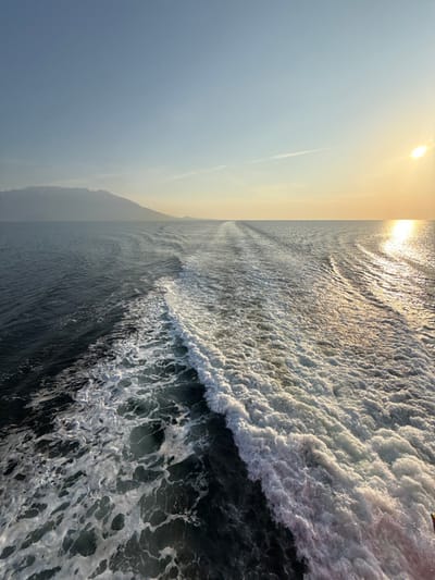 The image depicts a maritime scene observed from the stern of a moving vessel near Kato Karioutes, Greece, during sunset or late afternoon. A wide, turbulent wake of white foam dominates the lower half of the frame, trailing directly backward from the unseen vessel towards the horizon. The water to the left of the wake is dark blue-grey, while the water to the right, under the low-setting sun, exhibits a shimmering golden reflection. On the far left horizon, a distant, hazy mountain range or large island is visible. The sky transitions from a clear light blue on the upper left to a warm golden-yellow near the sun, positioned low on the far right. A faint, thin contrail or cloud streak is present in the upper-mid sky. No people, other vessels, or distinct man-made objects are identifiable within the scene.
