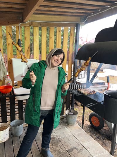 A woman in a green jacket and a light-colored hoodie is holding skewers of cooked chicken legs, standing on a wooden deck with a stone-paved area visible in the upper right corner of the image. She is near a barbecue grill with meat skewers cooking on it. In the background, there are wooden fences and trees. An orange bucket and bottles are visible near a white box on the deck. There are also a few metal buckets. The location is near Bol'shakino, Russia.