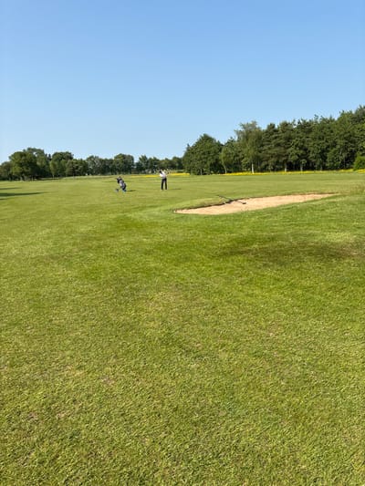 The image depicts a golf course, likely a sand trap, located in Pocklington, UK. The location is confirmed by the coordinates provided, 53.941 latitude and -0.761 longitude. The image captures a sunny day with clear skies. Two golfers are visible in the distance. One is standing on the green, while the other is in the sand trap. Both are dressed in light-colored clothing. The ground is covered in short, green grass, with a sand trap in the middle of the picture. The sand trap is located towards the far end of the course, with a line of trees in the background.