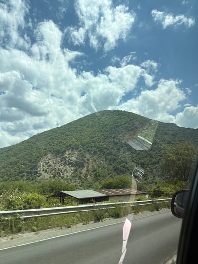 A landscape seen through the window of a car, likely an Audi, includes a view of a lush, green mountain, partially obscured by a cloudy blue sky. A road is visible on the right side of the image, next to a grassy roadside and a metal guardrail. A simple structure, possibly a small shed or shelter, is also visible near the roadside. The image provides no specific details to confirm it is taken in the city of Poleto, Bulgaria. The location cannot be definitively determined with the given information.