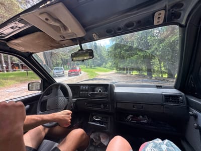 The image is taken from the interior of a car, looking forward through the windshield. The car appears to be driving on a dirt road surrounded by a dense forest of pine trees. In the distance, another car, a red hatchback, is visible on the road ahead, with a silver hatchback behind it. The car's interior shows a dashboard with an air conditioning vent, a radio, and a steering wheel. The driver's hands are visible on the steering wheel. The overall scene suggests a scenic drive through a rural or natural landscape. There is no indication of the city of Dedovo, Bulgaria.