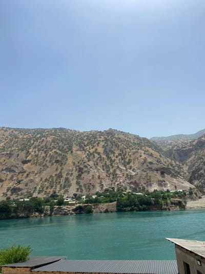 A wide, turquoise-colored river flows in the foreground, with ripples visible on its surface. On the far bank of the river, a small village is nestled among green trees. Houses with white roofs and some brown structures are visible. Behind the village, a steep, dry mountain range rises, covered in sparse vegetation and rocky outcrops. The sky above is clear and pale blue. In the lower foreground, two corrugated metal roofs of buildings are visible, one appearing to be part of a structure made of brick. The scene depicts a remote, mountainous area, possibly a rural village in Tajikistan.