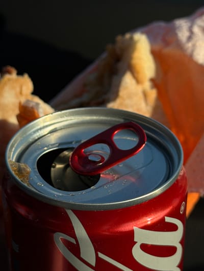 An opened red and silver soda can with a red tab is positioned in the foreground, slightly angled. A sandwich wrapped in orange paper is visible in the background. The sandwich's bread is a light beige. The background is a dark color, creating contrast with the brightly lit can and sandwich. Some condensation appears on the top of the can near the opening.