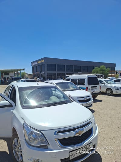 The image shows a daytime scene with several white cars parked on a dusty lot in front of a modern building. The building has large glass windows and signage that reads "LEGENDA SAWDA ORAVI" and "AVALON". Another sign on the building says "HÁJETXANA", which is Uzbek for "toilet" or "restroom".
The cars are mostly white, with one visible vehicle in the foreground that appears to be a Chevrolet. License plates are visible on some of the cars, with text such as "05 S 273 MA" and "95 R 564 PA". The sky is a clear blue, suggesting fair weather.
In the background, there are some trees and other buildings, indicating an urban or semi-urban environment. There are no people prominently featured in the image, but some figures are visible near the building in the distance.
A timestamp is overlaid on the image, reading "23 июня 2026 г. 12:33", which translates to "June 23, 2026, 12:33". Another text overlay reads "952332 Galaxy", possibly referring to a phone model. The scene appears to be a car dealership or a parking area near commercial establishments. The overall impression is a bright, sunlit day in an area with a mix of modern architecture and parked vehicles.