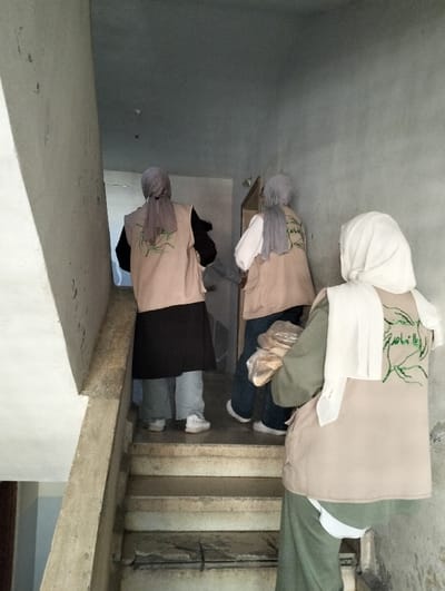 Three individuals, presumably adult females, are depicted from behind, ascending a worn concrete staircase within a building in قرية الضبية, Syria. All wear light-colored vests featuring green embroidered designs and Arabic script on the back. The individual in the foreground, wearing a white hijab and an olive-green garment under her vest, carries a light-colored, possibly plastic-wrapped package in her right hand. The second person, positioned mid-stairs, wears a light gray hijab, a white top, and blue jeans under her vest. The third person, further up the stairs, wears a light gray hijab, a black top, light blue jeans, and white sneakers. The staircase and surrounding walls are unpainted concrete or plaster, showing signs of wear and minor damage. A doorway is visible at the top of the visible stair section. The scene suggests a distribution or delivery activity.