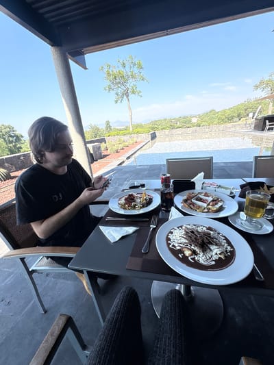A man sits at an outdoor table in Pueblo Nuevo de Jasso, Mexico, looking at his phone. The table is set with plates of food, including what appears to be enchiladas, drinks, and condiments. The background shows a sunny day and a landscape of trees and buildings.