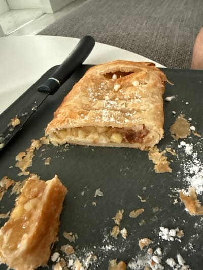 An apple strudel lies on a dark grey rectangular plate next to a black-handled knife. The strudel is golden brown and dusted with powdered sugar. Some crumbs and powdered sugar are scattered on the plate around the strudel, and a small piece of strudel has been cut off and is visible in the lower left corner of the image. The background of the image is blurred, showing a white table and a grey sofa. The scene appears to be indoors, likely in a home environment in Gothenburg, Sweden. There are no people visible in the image.