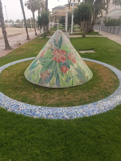 The image depicts a conical sculpture painted with floral designs, situated in a grassy area bordered by a mosaic tile planter. The sculpture is located near the Águilas promenade, overlooking the beach. Palm trees and other vegetation are visible in the background, along with buildings and a clear sky. A person is walking a dog on the promenade in the distance. The overall scene suggests a public park or recreational area in a coastal city.